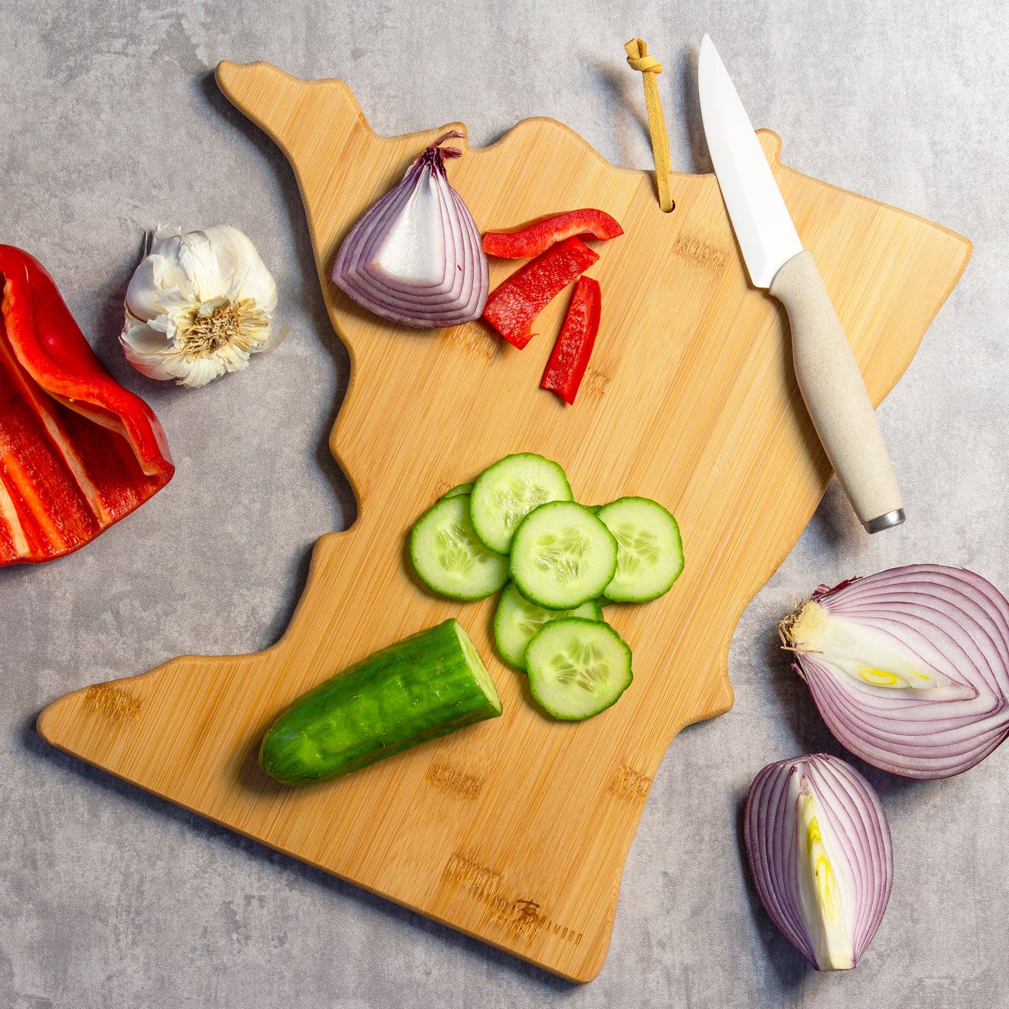 The Minnesota Cutting Board with Artwork by Fish Kiss™, crafted from bamboo and shaped like the state of Minnesota, holds sliced cucumber and bell pepper, with a knife resting on top. Surrounding it are garlic, sliced onion, and additional bell pepper on a sleek gray surface.