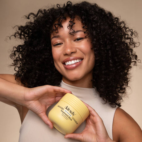 Smiling person with curly hair holds a yellow jar of Kitsch Hair: Moisturizing Coconut Oil Deep Conditioning Treatment Mask 10 oz, against a neutral background.