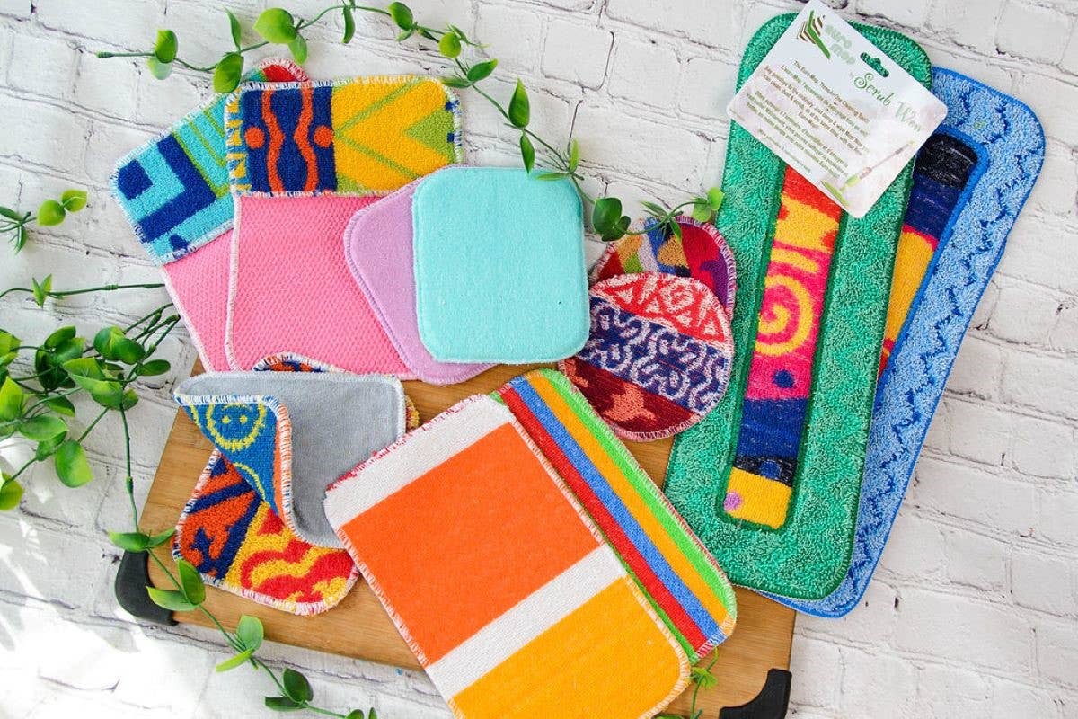 Scrub: Kitchen Scrubber Scrub-Wow reusable cleaning cloths and multipurpose scrubby pads are arranged on a wooden board with green vines against a white brick background.
