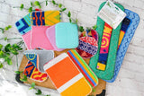 Scrub: Kitchen Scrubber Scrub-Wow reusable cleaning cloths and multipurpose scrubby pads are arranged on a wooden board with green vines against a white brick background.