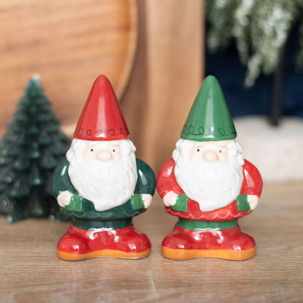 Two Shakers: Gnome Salt & Pepper Shakers, small ceramic figurines featuring red and green hats and suits, standing on a wooden surface near a tiny pine tree decoration, offer unique home decor that's perfect for those seeking winter forest-inspired giftware.