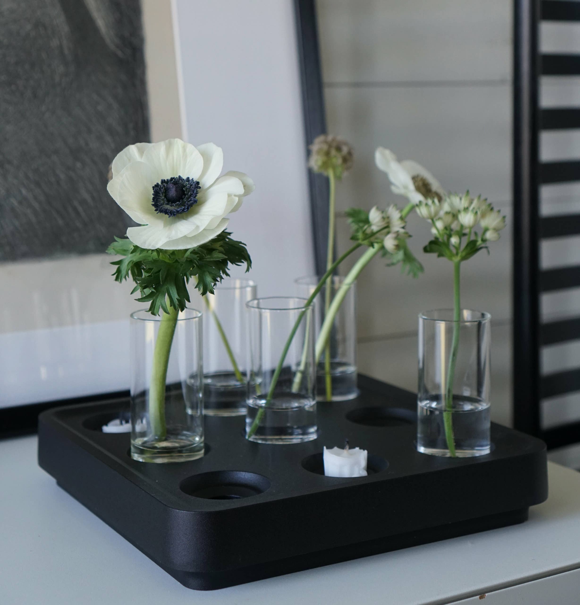 Mini vases filled with white flowers are elegantly arranged on a black tray, paired with a candle holder from the Mini Vases pack of 4, and complemented by a framed picture in the background.