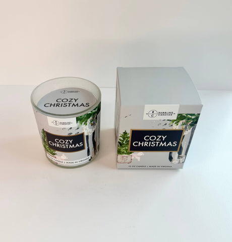 The 12 oz Cozy Christmas Candle sits beside its matching box, both featuring festive holiday designs and fragrance, arranged on a white surface.