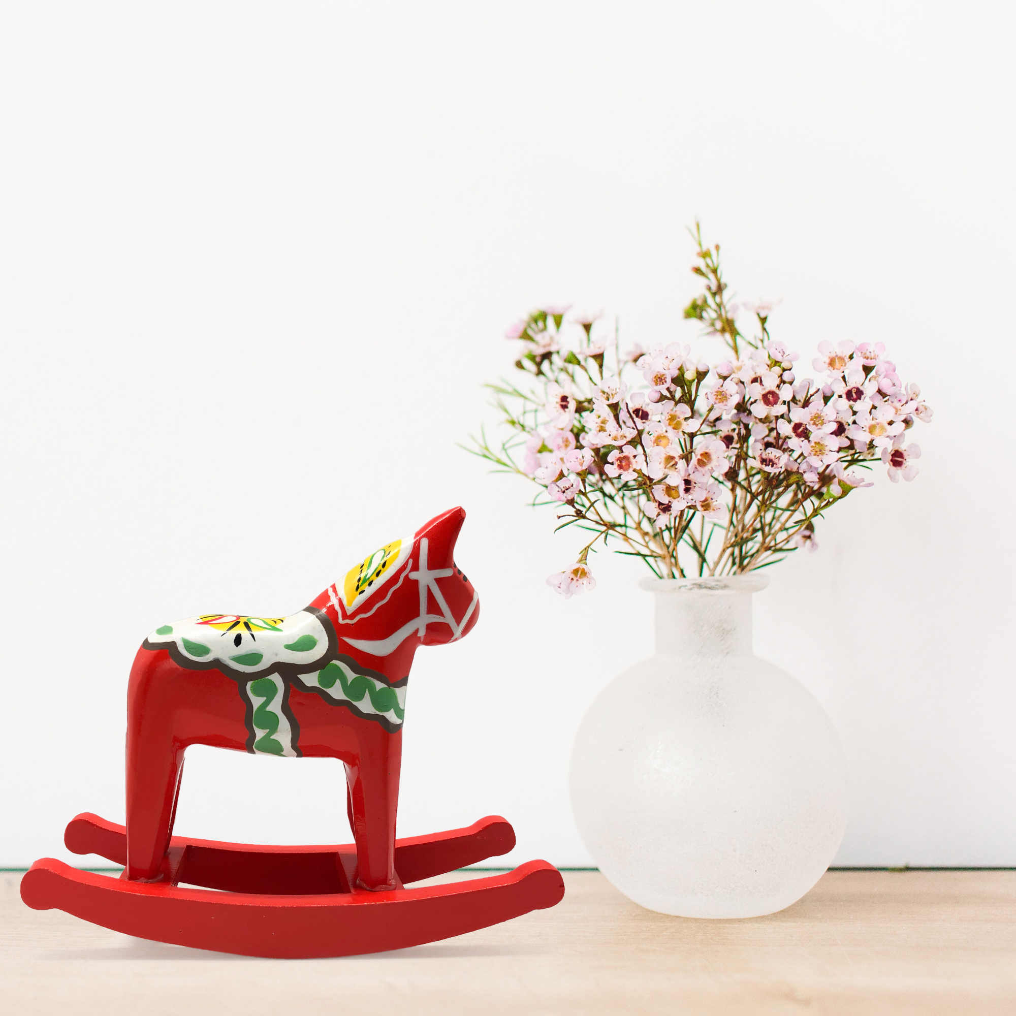 A red Dala Rocking Horse figurine with multi-colored kurbits, embodies Scandinavian motifs beside a frosted glass vase holding pink flowers on a wooden surface, subtly showcasing Swedish heritage.