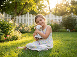 A young girl in a floral dress sits on green grass, smiling and hugging her Faire - The Petting Zoo Plush: Goat 9″ Tall Stuffed Animal, with sunlight and a white picket fence in the background.