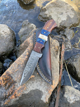 A Handmade High Carbon Damascus Steel Hunting Knife by Titan International, featuring a patterned blade, diamond wood and stag horn handle, and leather sheath, rests on a rock near a stream with water and rocks in the background.