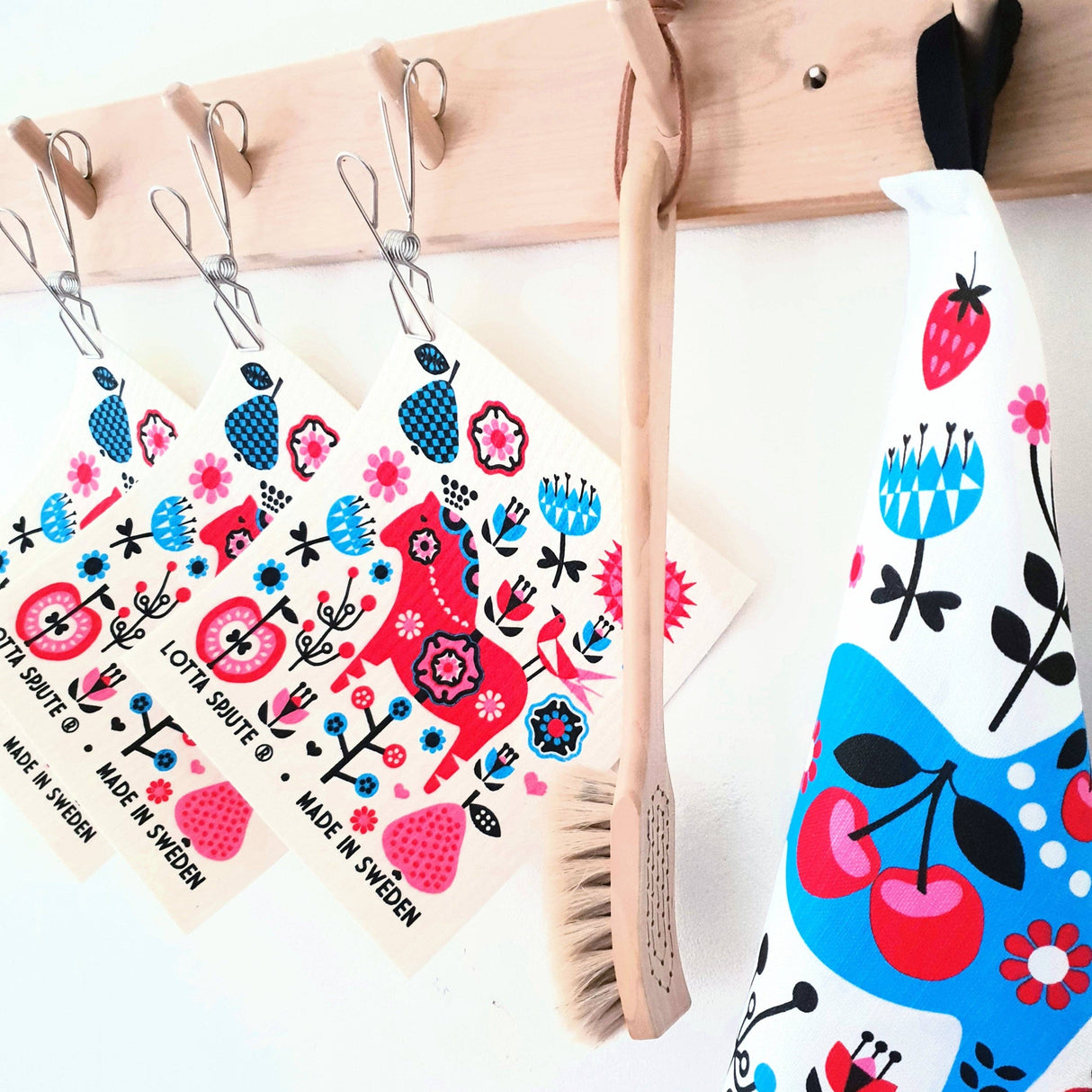Wooden rack with three eco-friendly Swedish dishcloths in red and blue floral patterns, a wooden brush, and a Tea Towel: Swedish Dala Horse Linen - Cotton Kitchen Towel Lotta Spjute Sweden with colorful designs hanging on hooks.