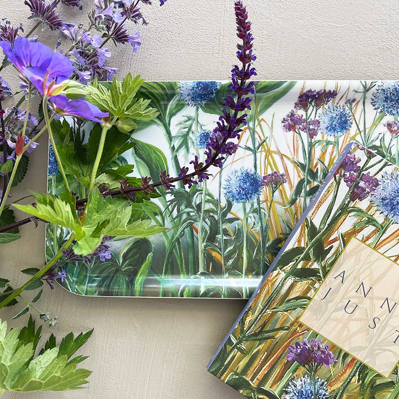 The Koustrupco Anne Just Birch Veneer Tray (32x15 cm), adorned with beautiful blue and purple flowers, rests on a textured surface alongside a book and sprigs of purple and green flowers, capturing the artistic essence of Anne Justs botanical creations.