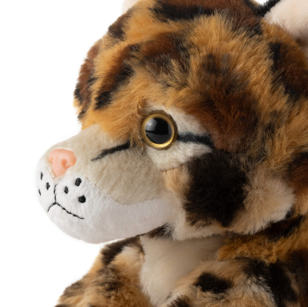 Close-up of a plush toy cat with brown, black, and white fur, golden eyes, pink nose, visible whisker spots, facing slightly left.