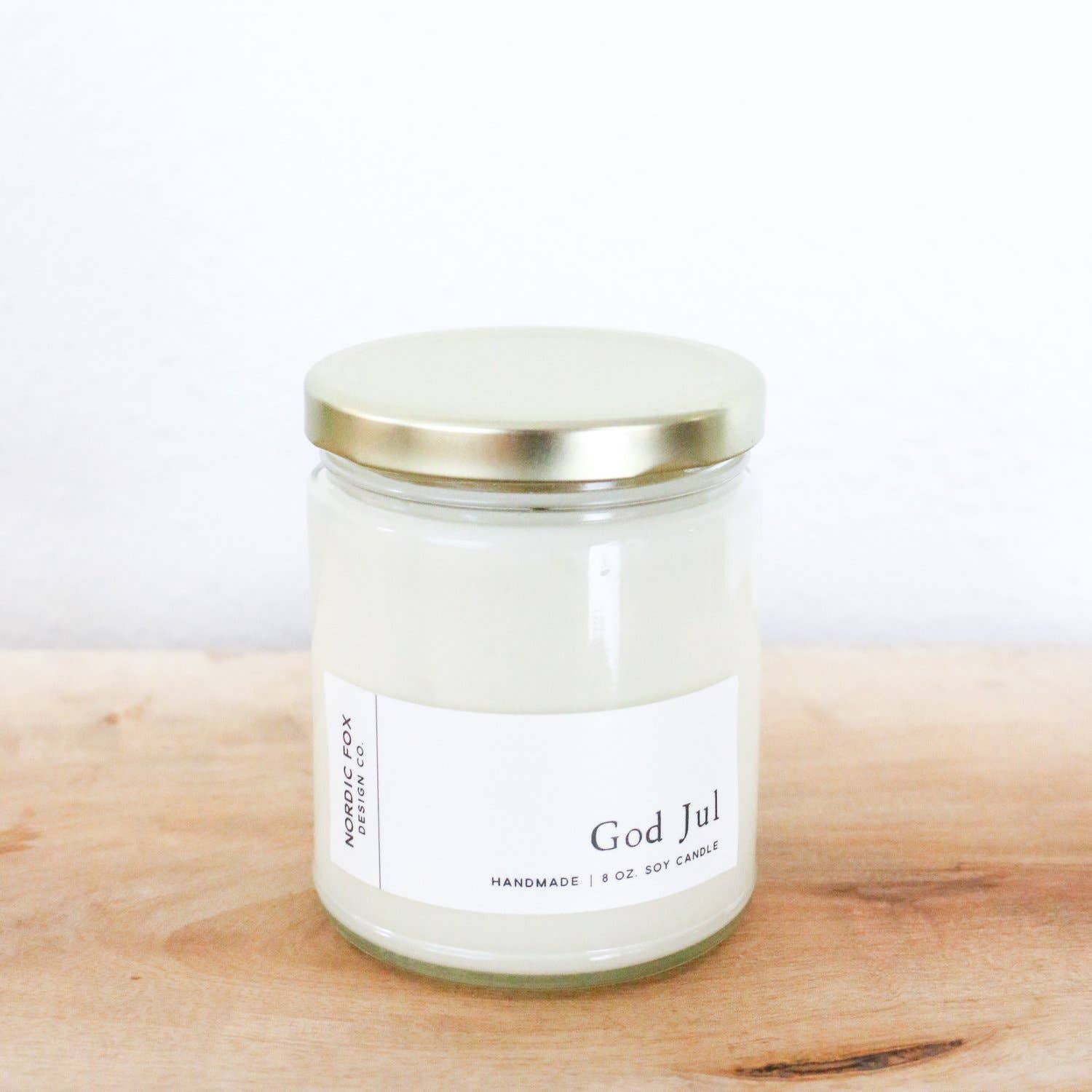 A white Candle: God Jul Handmade Soy Candle sits on a wooden surface in a clear glass jar with a gold lid. The 8 oz. jar candle is a festive Nordic holiday decor piece, ideal as a handmade Christmas gift or seasonal accent.