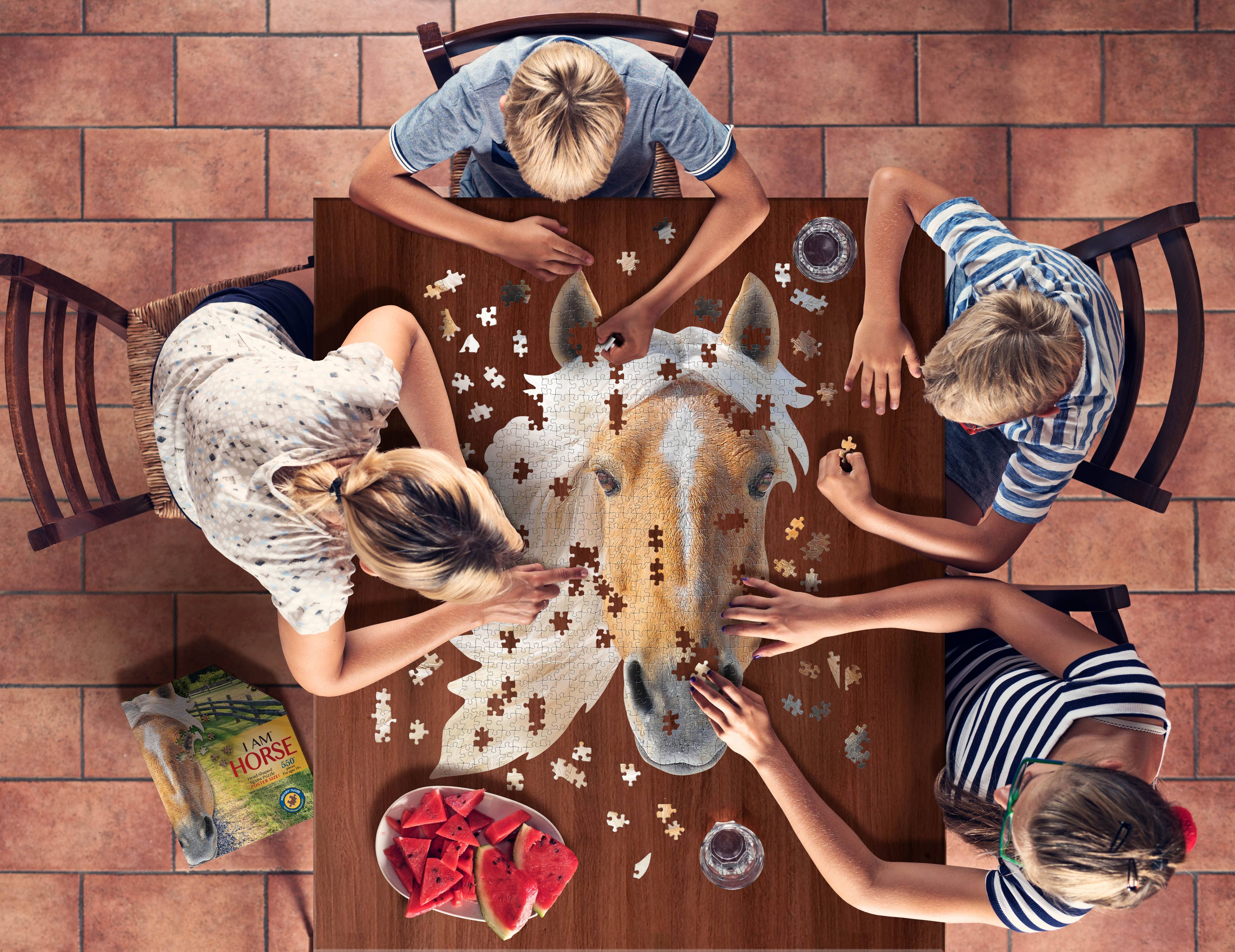 Four people are gathered around a wooden table, working on the "I AM Horse" 550-piece educational jigsaw puzzle. The scene is complemented by a book about horses and slices of watermelon, enhancing the atmosphere.