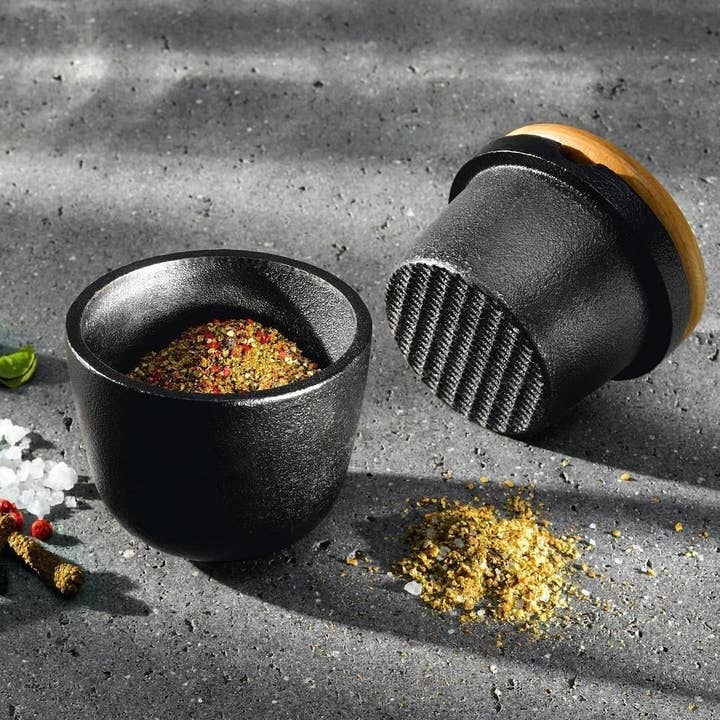 The Skeppshult Swing Spice Mill with Walnut Lid in black cast iron holds mixed spices, while ground spices and salt are scattered on a gray surface.
