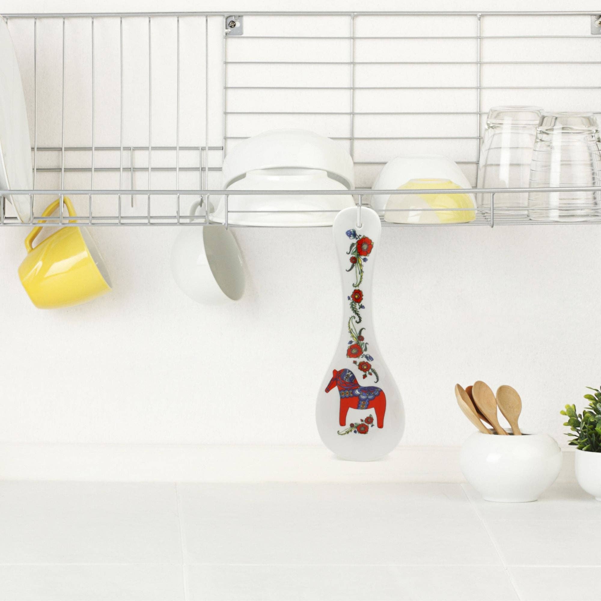 A kitchen exuding Scandinavian elegance features a wire rack with cups, glasses, and dishes. Below, the Spoon Rest: Swedish Red Dalarna Horse Ceramic adds a vivid touch to a counter adorned with bowls, wooden spoons, and a small plant.