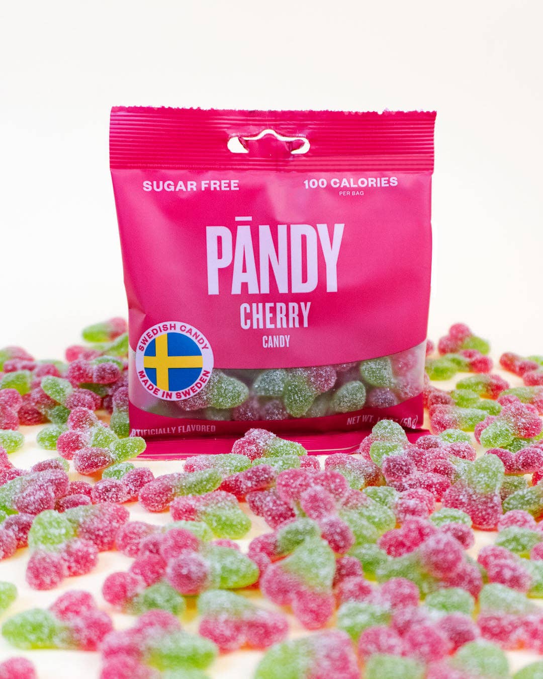 A bag of Pandy's Sugar Free Sour Cherry candy, weighing 1.8 ounces, with a delightful sourish flavor is displayed on a white surface, surrounded by scattered red and green gummy bears. The Swedish candy label is clearly visible on the packaging.