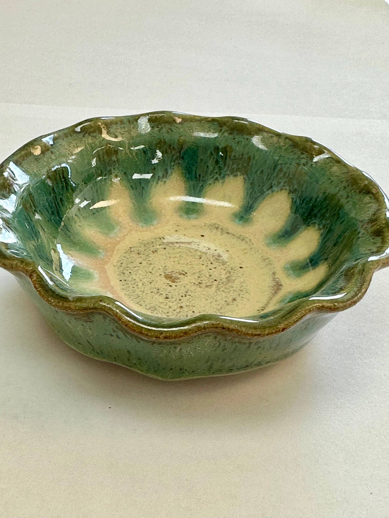 A small handmade stoneware bowl made of glass and clay (4 3/8" diameter) with a green scalloped edge and mottled cream interior, shown on a white surface.