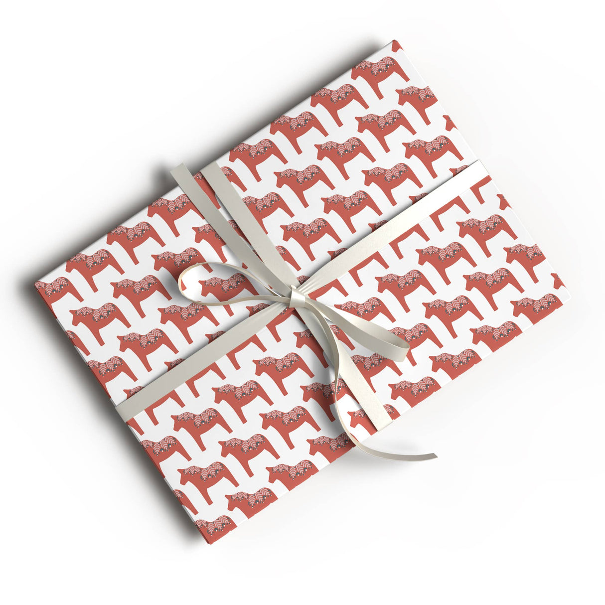 A rectangular gift box wrapped in Juniper Blue's Swedish Dala Horse Gift Wrap (glossy, 2 sheets, 20"x29" each) tied with a white ribbon and bow, displays charming Scandinavian style on a white surface.