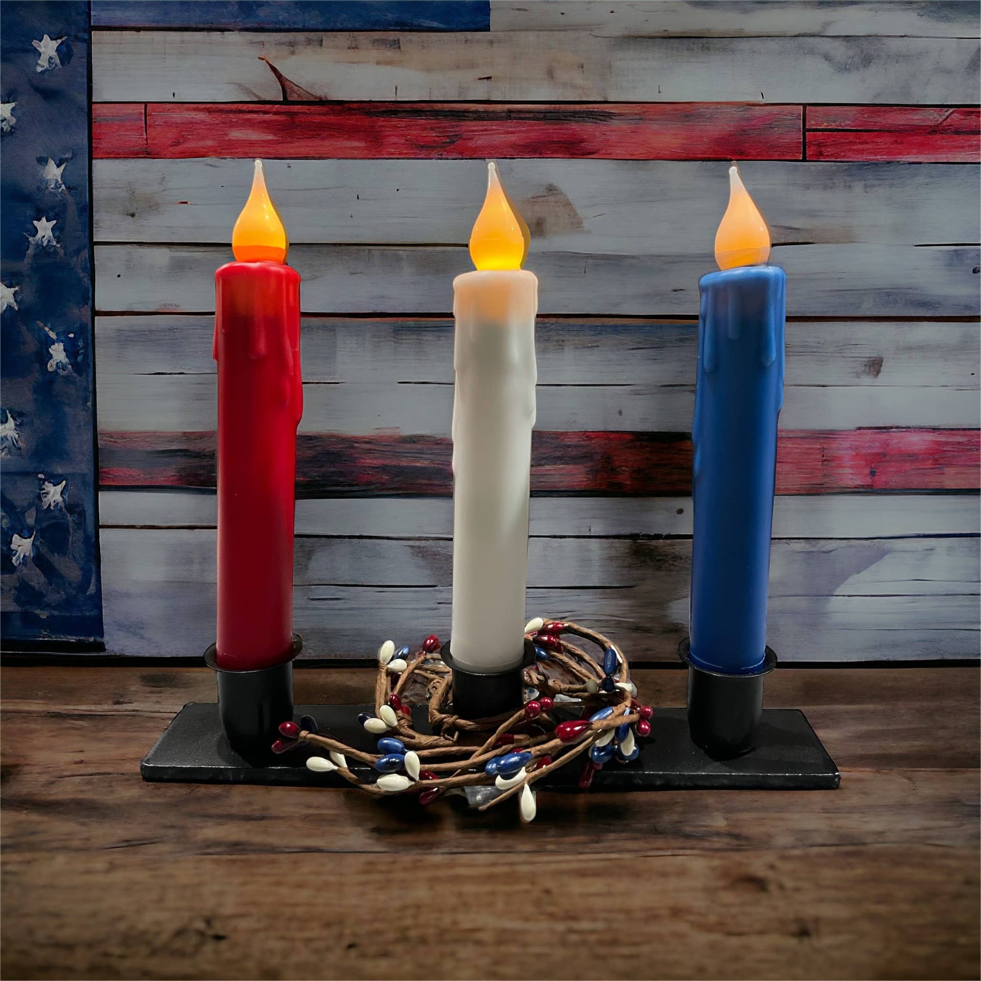 Three solid white LED battery-operated timer taper candles, each measuring 7 inches, are elegantly placed on a stand. A small decorative wreath with berries encircles the base, while a realistic flickering LED bulb adds charm against the American flag-themed wooden background.