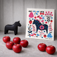 A Swedish Dala Horse dish cloth by artist Lotta Spjute, featuring a colorful floral horse design, is displayed on a gray surface with seven red apples and a wooden panel background.