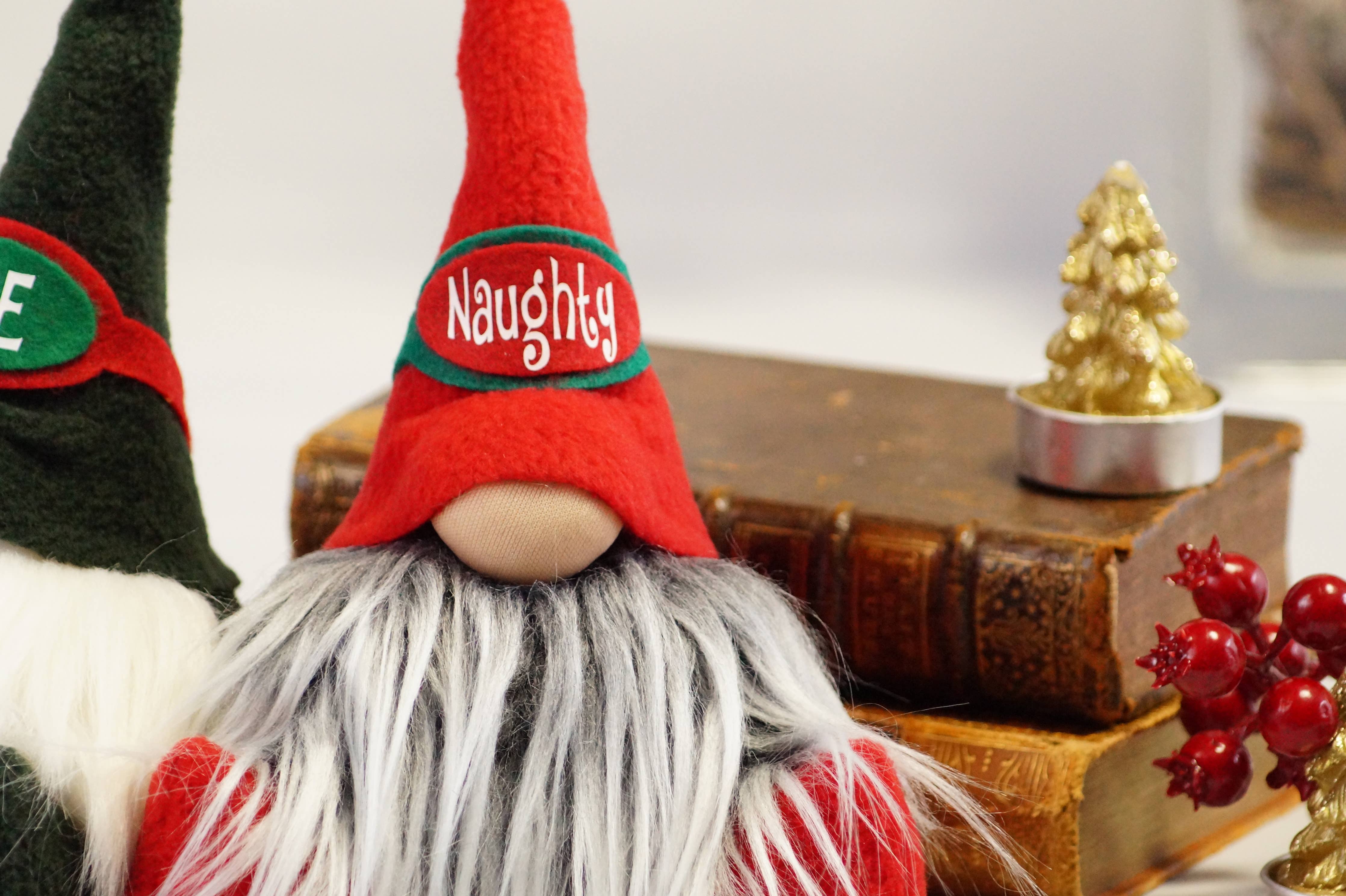 The handmade Naughty Christmas Gnome, known for its playful expression and red hat, is positioned next to two vintage books, a gold tree ornament, and vibrant red berries.