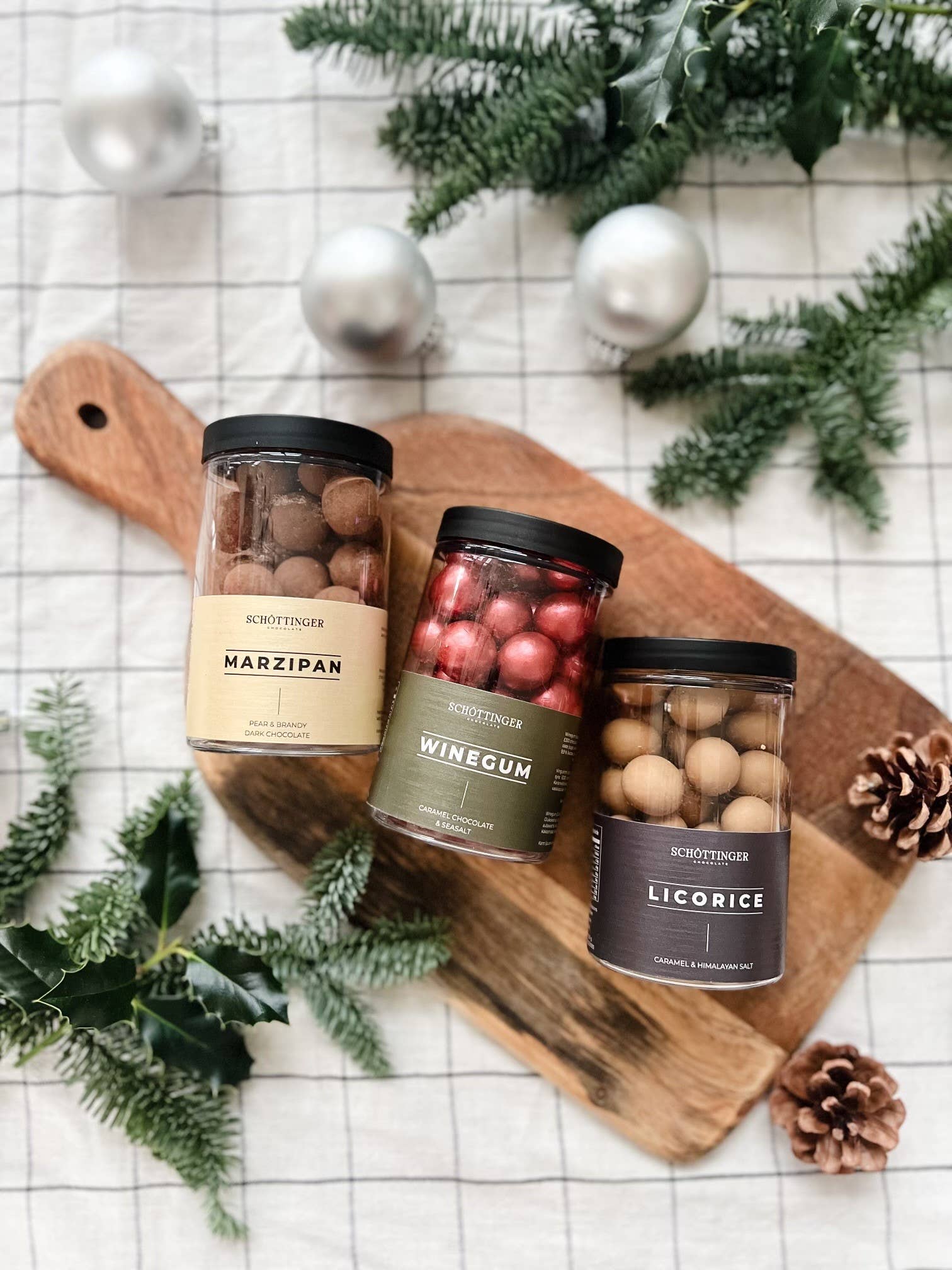 A jar labeled "Candy: Classic Winegum Seasalt & Caramel Chocolate 250 g" sits on a wooden board adorned with red amber pine cones and greenery, all set atop a checkered cloth.