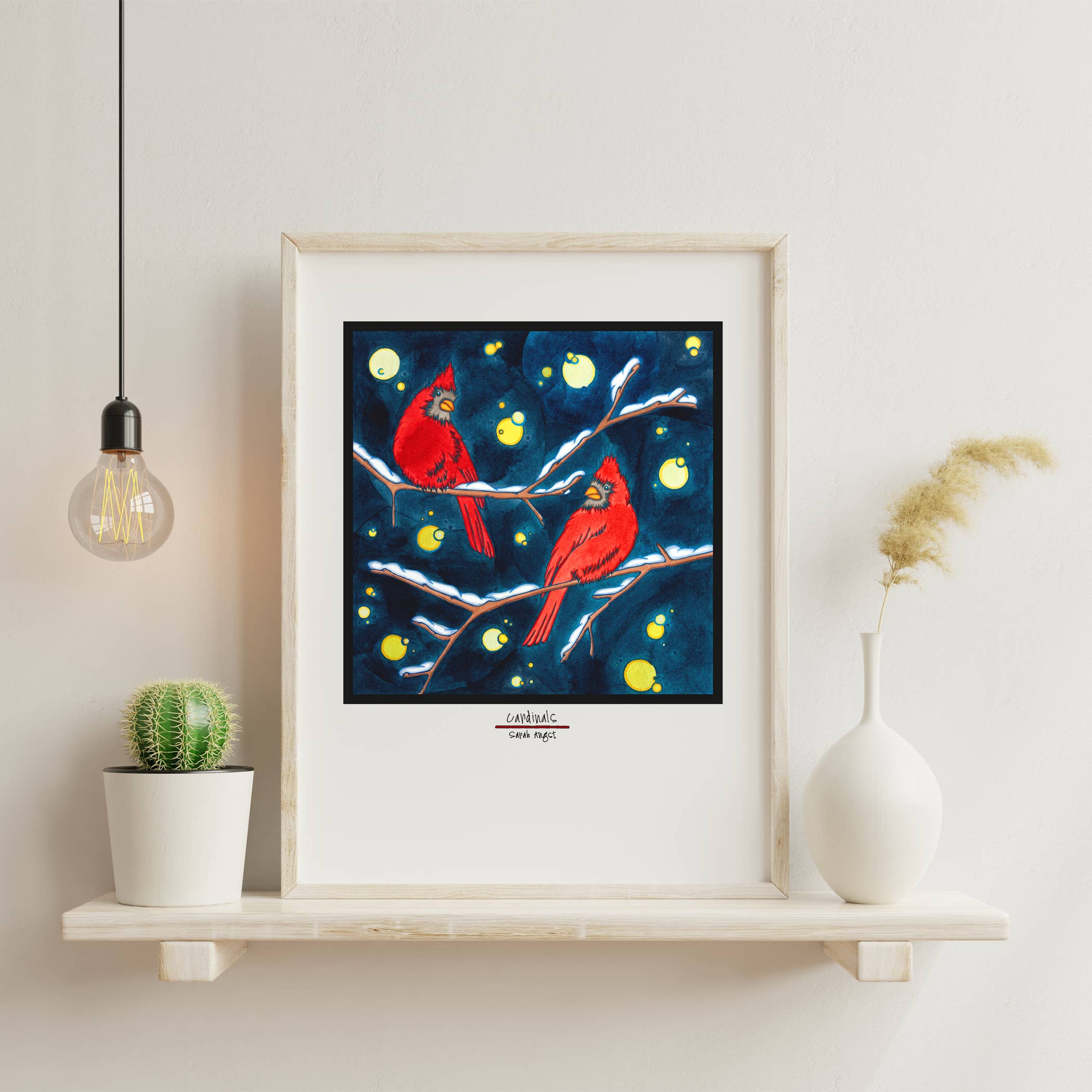 The "Artwork: Winter Cardinals" 8"x10" Giclee Print depicts two cardinals perched on branches against a dark blue background with light spots, elegantly displayed on a white shelf alongside a cactus, vase, and hanging bulb—a perfect addition to your Christmas decor.