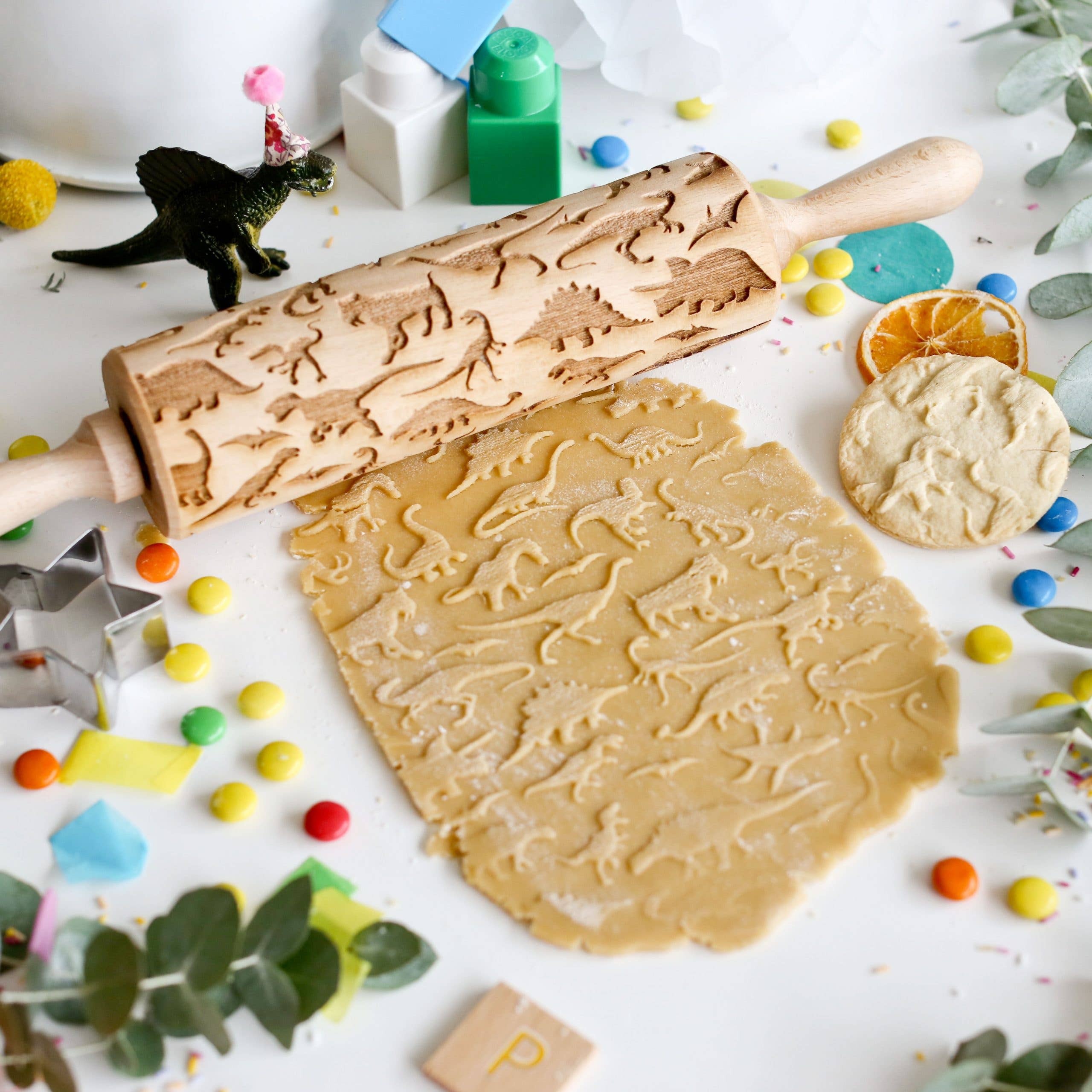 The Dinosaur 1 Embossing Rolling Pin, a handmade piece with laser-engraved dinosaur designs, embosses cookie dough on a table adorned with leaves, colorful candies, cookie cutters, and decorative items.