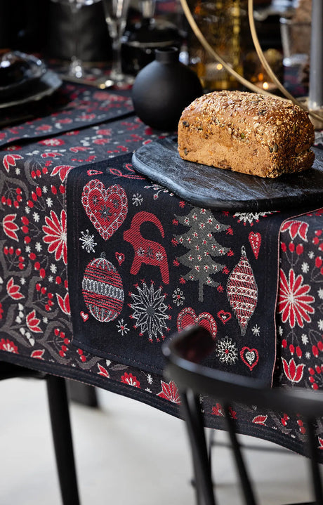 Black tea towel with a red, white, and grey pattern of hearts, Dala horses, and flowers, shown under a loaf of seeded bread on a black board.