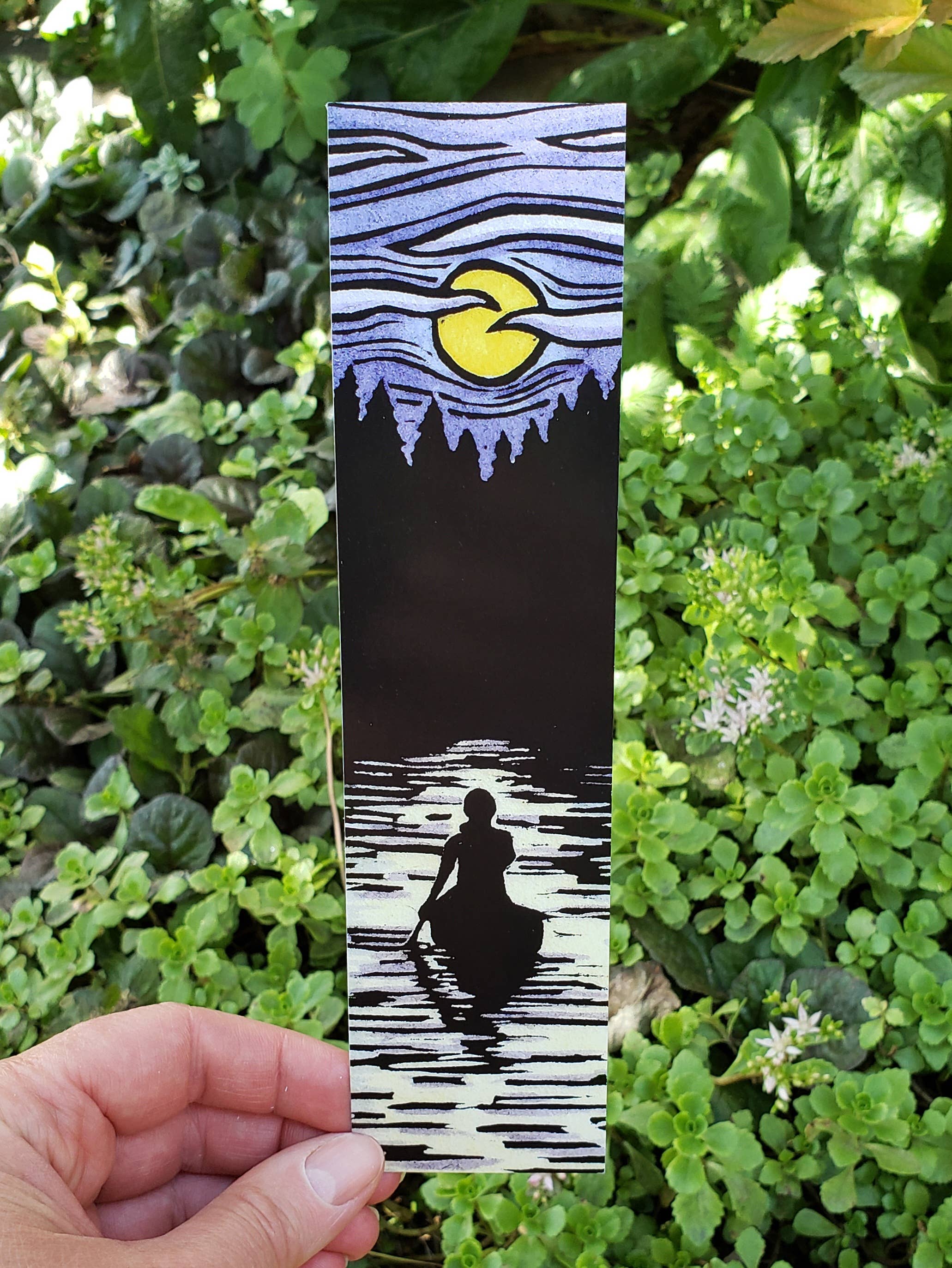 Hand holds the Solitude Bookmark, showcasing art of a canoeist under a yellow moon and rippling sky, encircled by greenery. A stunning landscape embodying Sarah Angst Art.