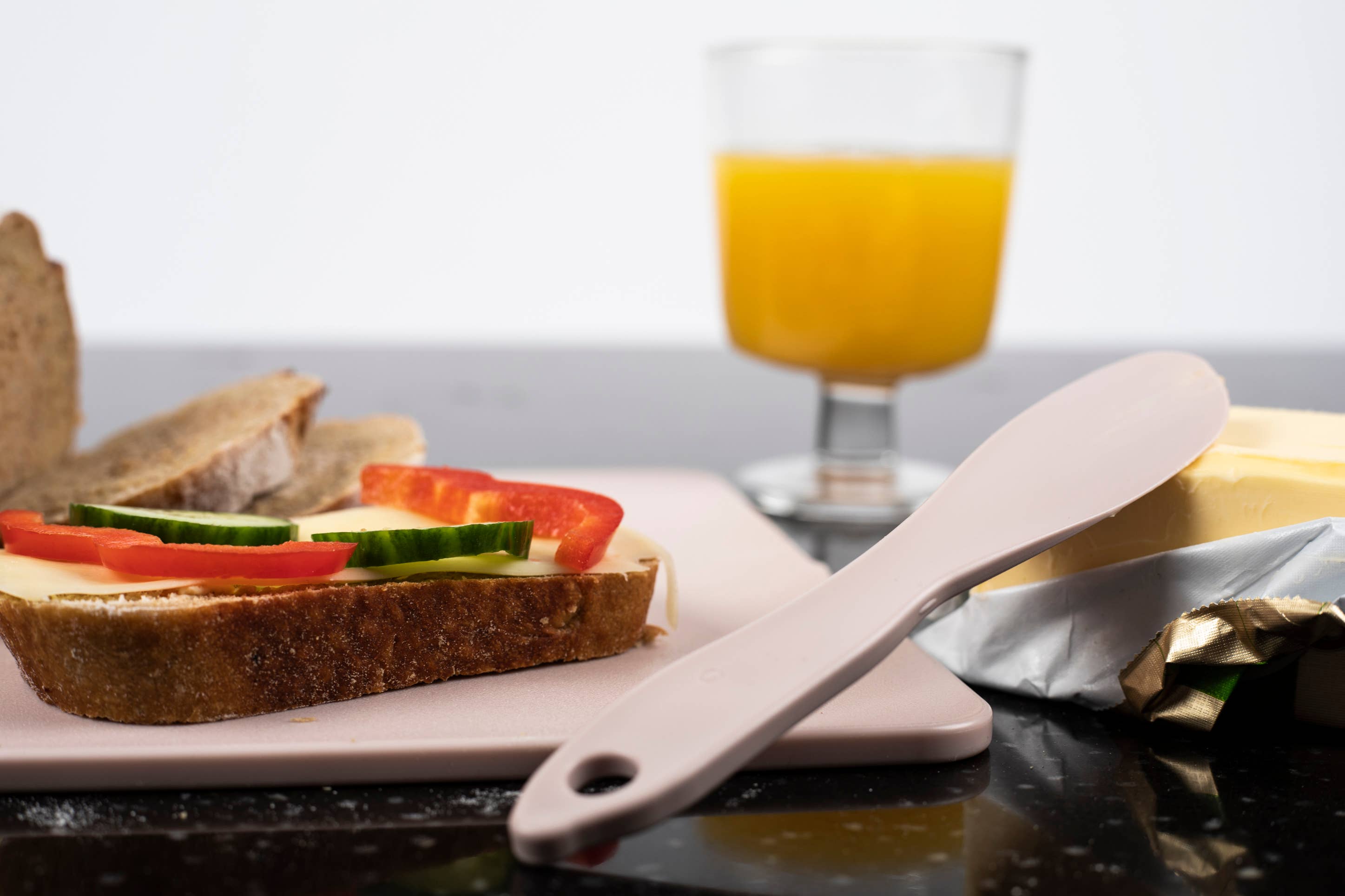 A slice of bread with cheese, cucumber, and bell pepper sits on a sustainable cutting board, accompanied by a Swedish Natural Single Spreader in beige alongside rich butter, while a refreshing glass of orange juice completes the scene.