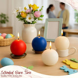 Four Dusty Rose Ball Candles, each 2.5" in diameter, are burning on a table next to a vase of flowers, a picture frame, and Easter decorations, with two people blurred in the background.