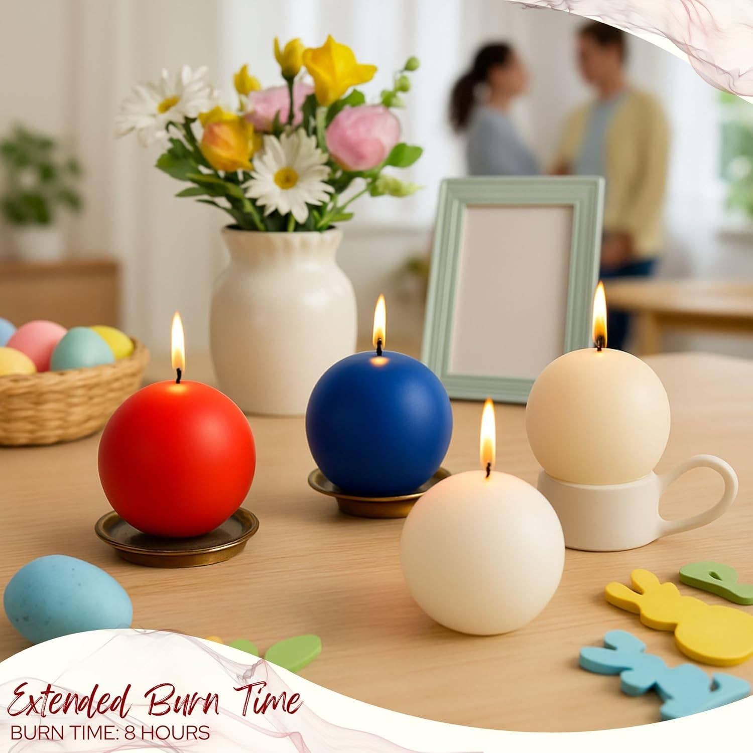 Four Champagne Ball Candles, 2.5" diameter—red, blue, and two white unscented—burn on a table with a vase of flowers, photo frame, and decor. Two people are blurred in the background.