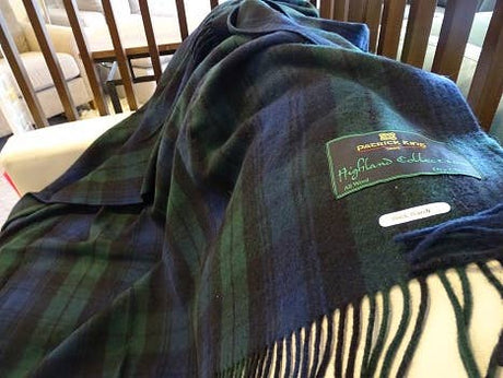 A Black Watch Highland Tartan Plaid Wool Blanket (69"x62") in dark green and navy, with fringed edges, is draped over a white surface. The label reads Patrick King Highlands Collection.