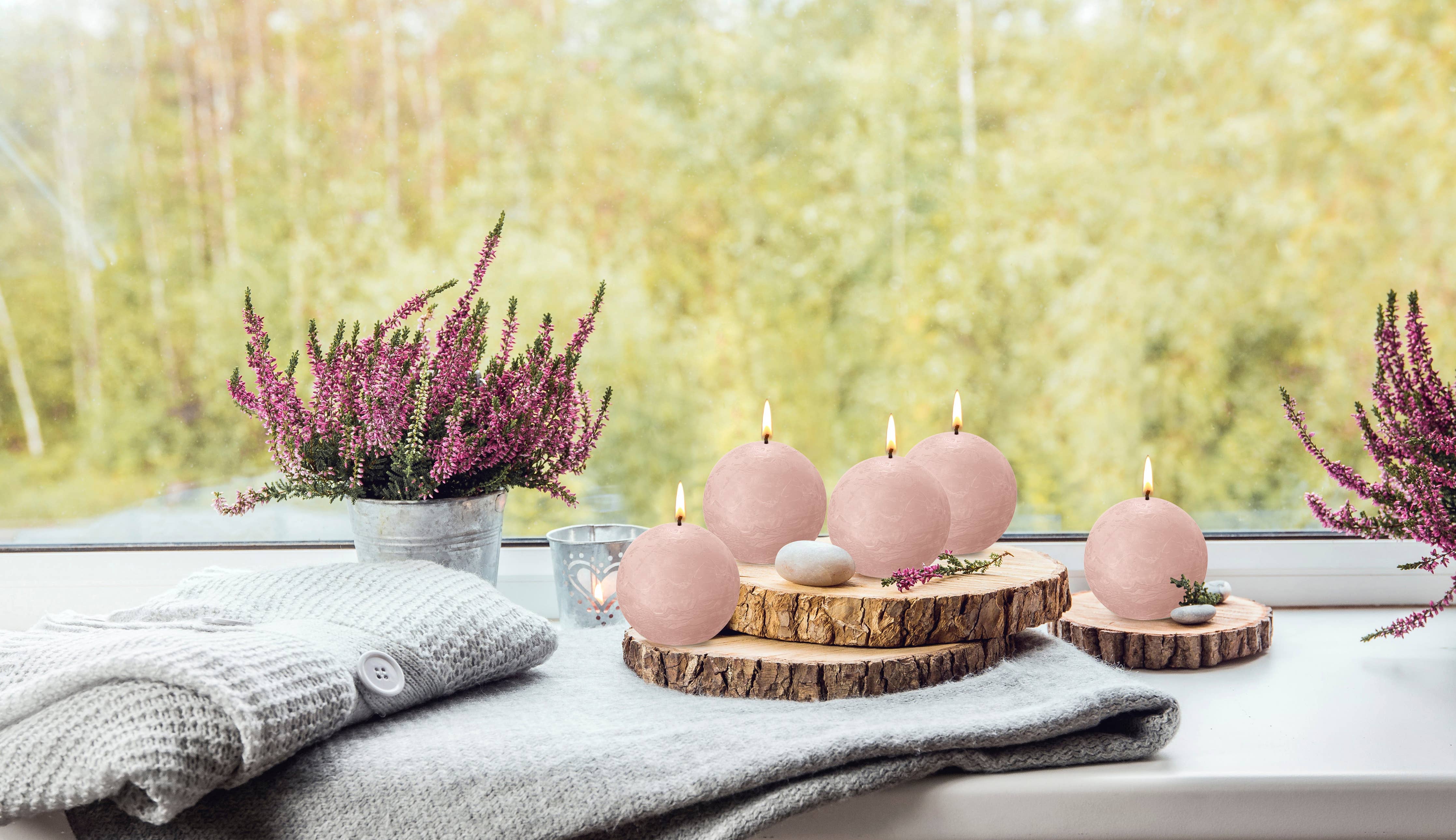 A Misty Pink Rustic 3" Ball Candle is lit on a wooden slice next to a potted purple heather and folded knit blankets on a windowsill, with a blurred outdoor view in the background.