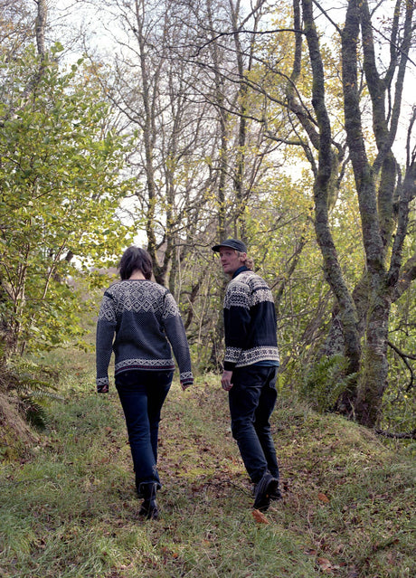 Two people walk on a grassy path through wooded trees, one wearing the Stavanger Norwegian Wool Cardigan in Black and looking back toward the camera.