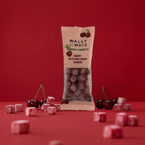 A 2 oz packet of Wally & Whiz US Danish Sweets Cherry with Sour Cherry Vegan Gummies stands upright on a red surface, surrounded by scattered gummies and fresh cherries against a solid red background.
