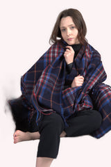 A woman sits on the floor with bare feet, wrapped in the Esprit du Quebec Deluxe Highland Blanket, wearing a black sweater and pants against a light background.