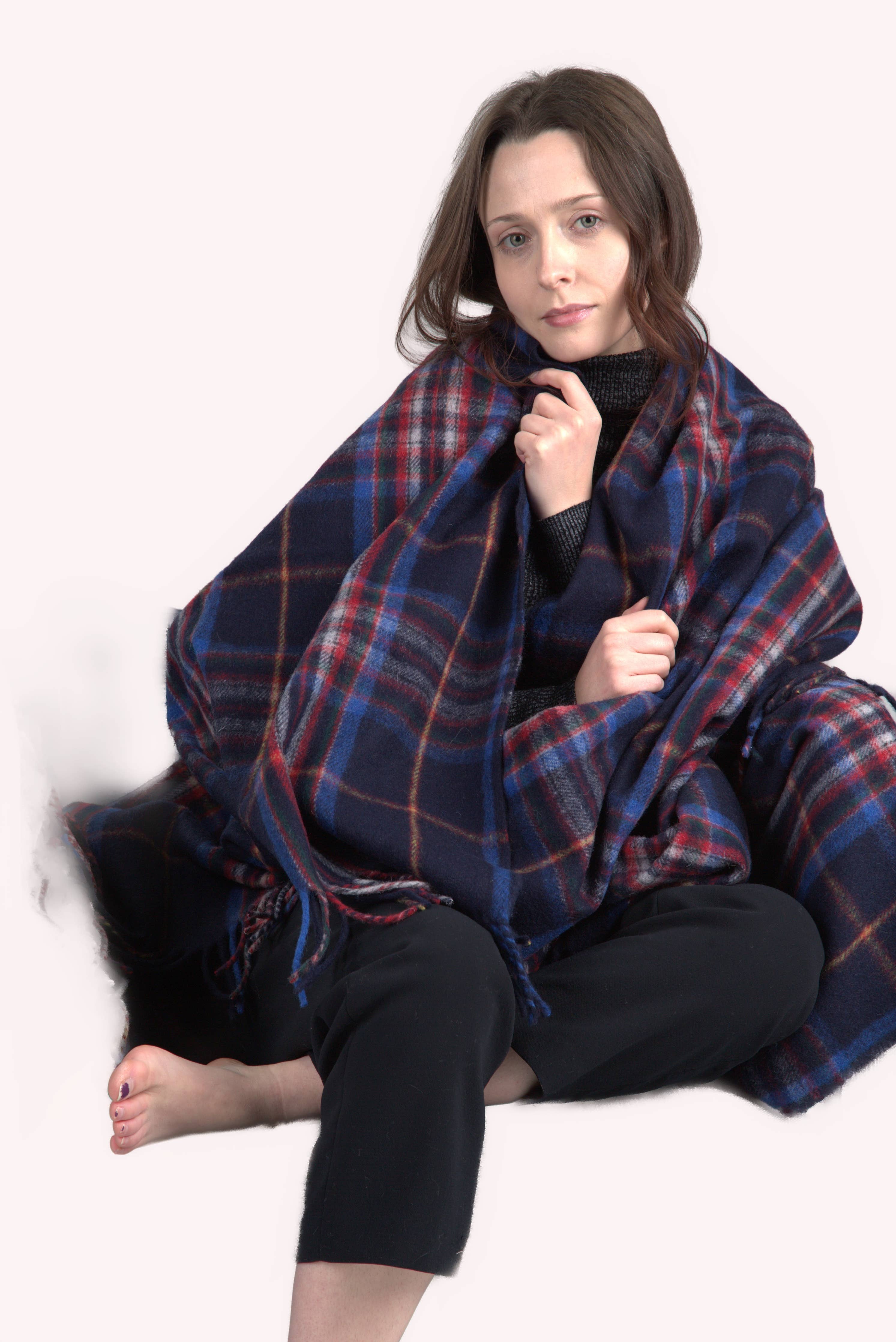 A woman sits on the floor with bare feet, wrapped in the Esprit du Quebec Deluxe Highland Blanket, wearing a black sweater and pants against a light background.
