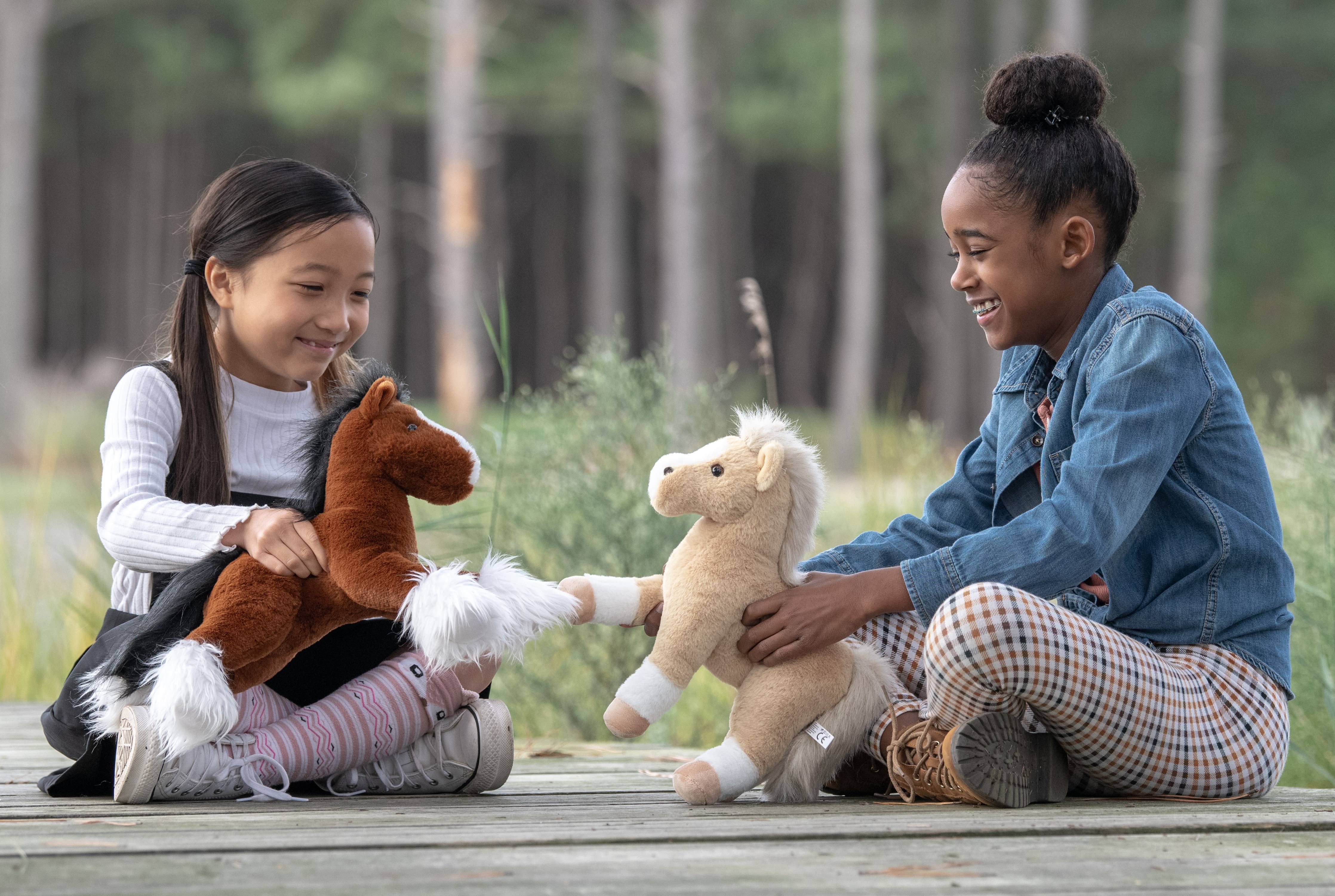 Two girls sitting on a wooden platform, each holding a "Plush: Horse Black w/ Star Standing 12"", smiling at each other in an outdoor setting.