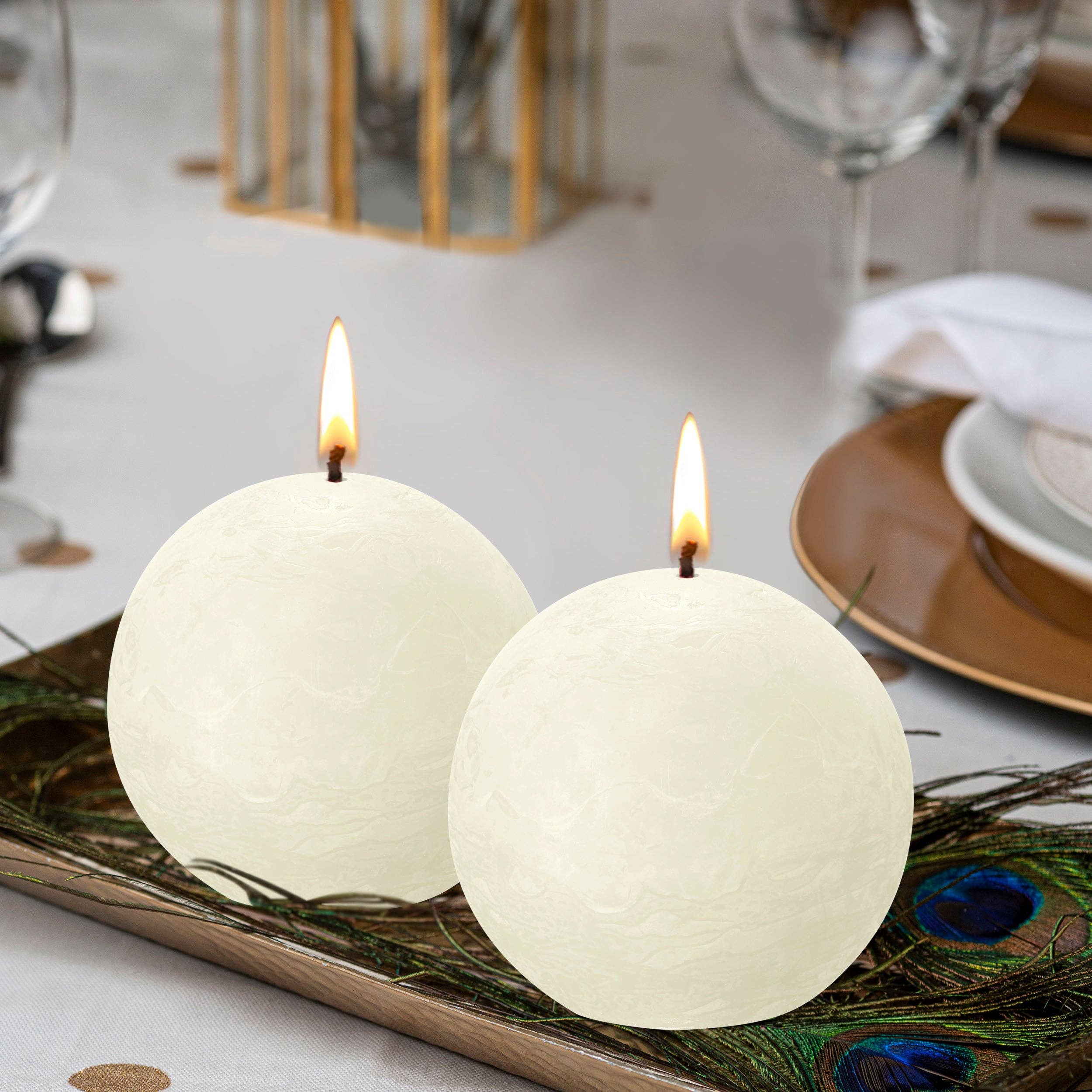 Two Ash Rose Rustic 3" Ball Candles, made from eco-friendly, natural plant-based wax, lit on a decorative tray with peacock feathers, set elegantly on a dining table.