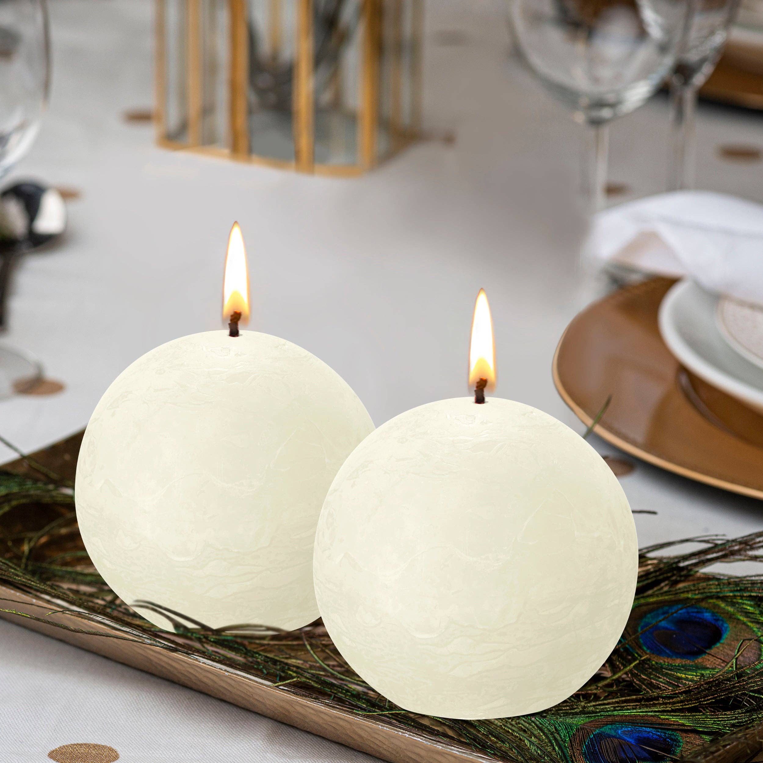 An Ivory Rustic 3" Ball Candle rests with a soft flame on a decorative tray with peacock feathers, elegantly arranged among plates and glasses on the dining table.