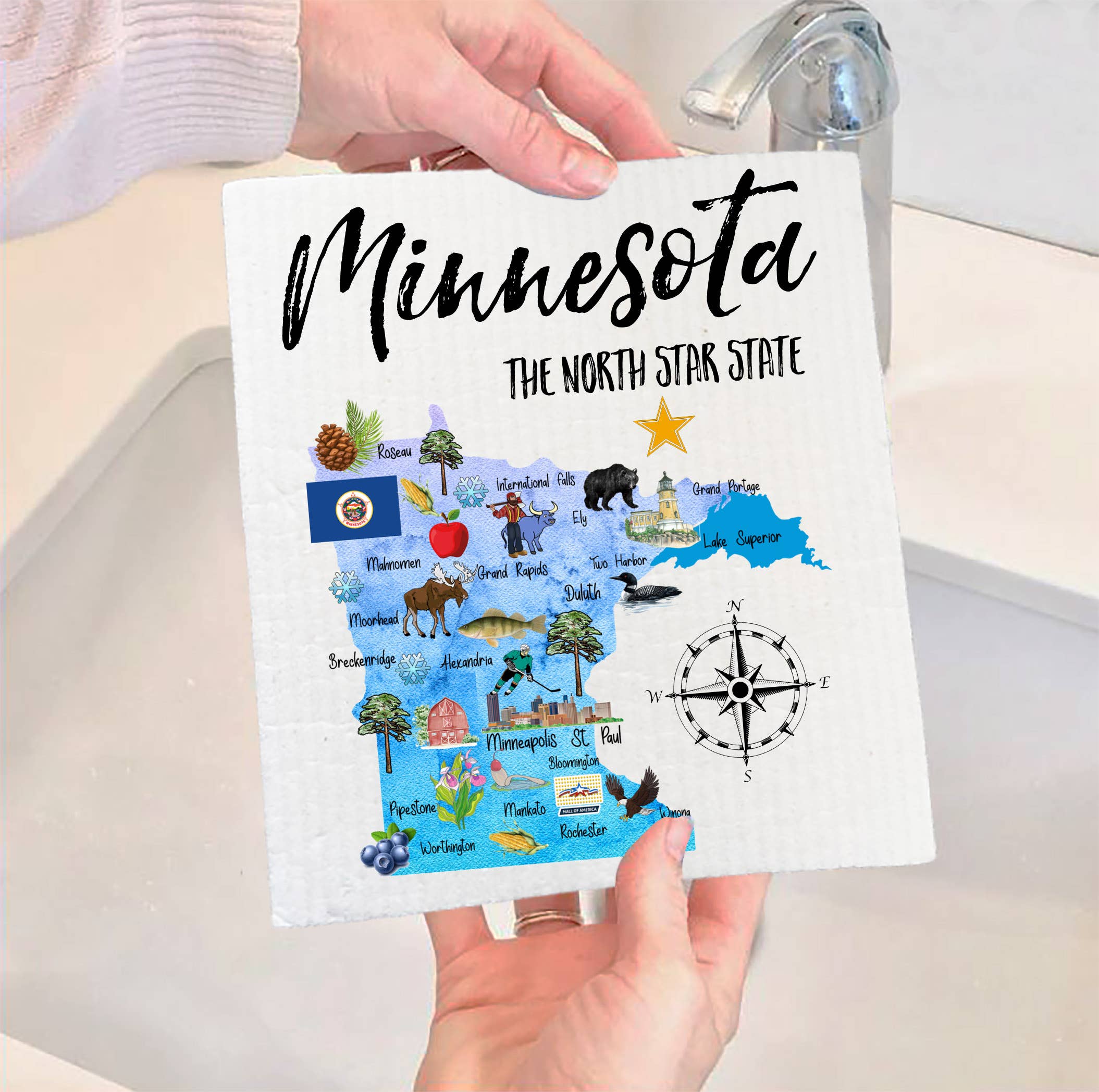 A person holds the Dish Cloth: Minnesota State Map Souvenir SWEDISH DISH CLOTH near a sink, featuring a vibrant Minnesota map design with state symbols, landmarks, and the phrase "Minnesota The North Star State." This eco-friendly dishcloth highlights state pride while being compostable and biodegradable.