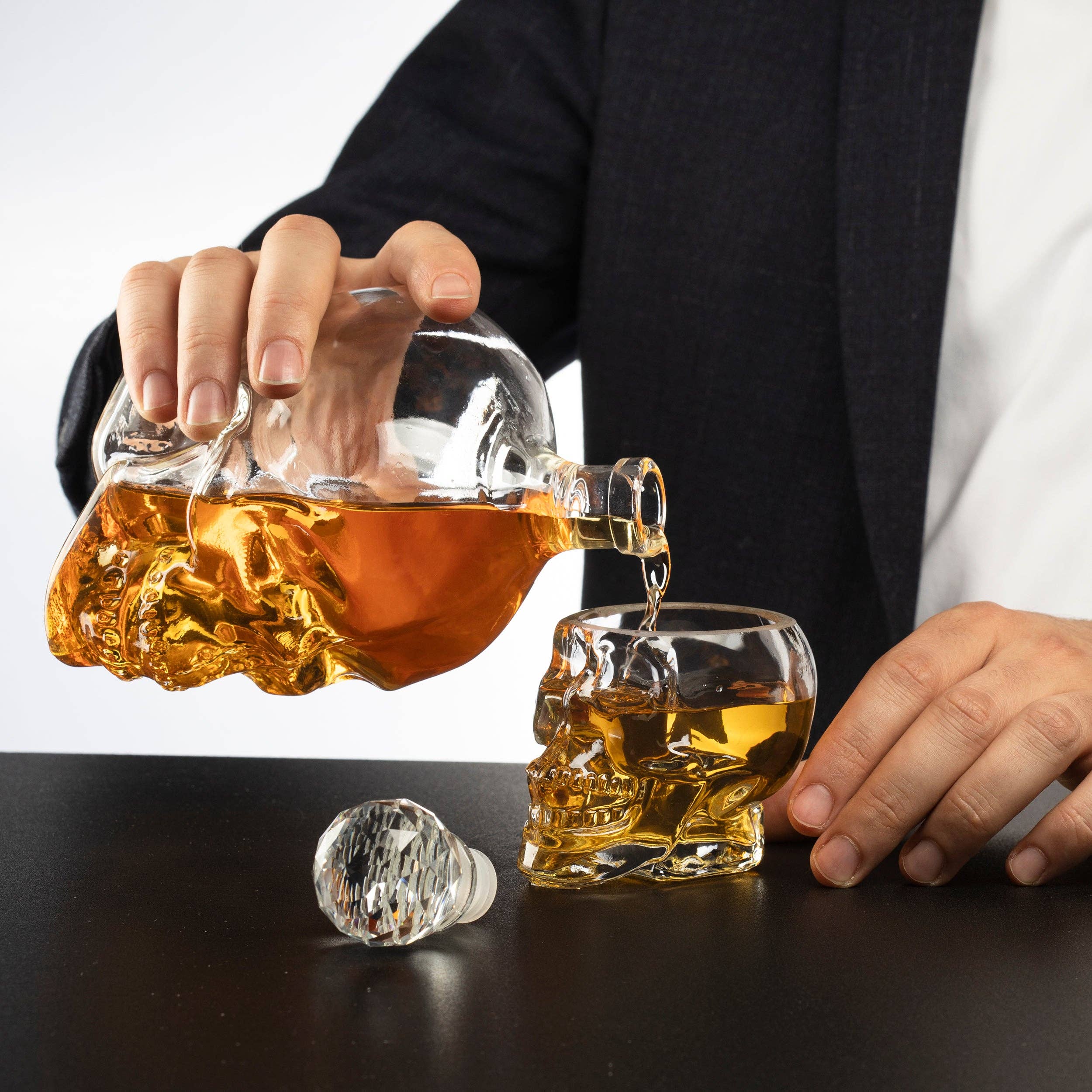 At a pirate-themed party, someone pours amber liquid from the Skull Decanter Set with 4 Skeleton Skull Shot Glasses on a black table, while a crystal stopper rests nearby.
