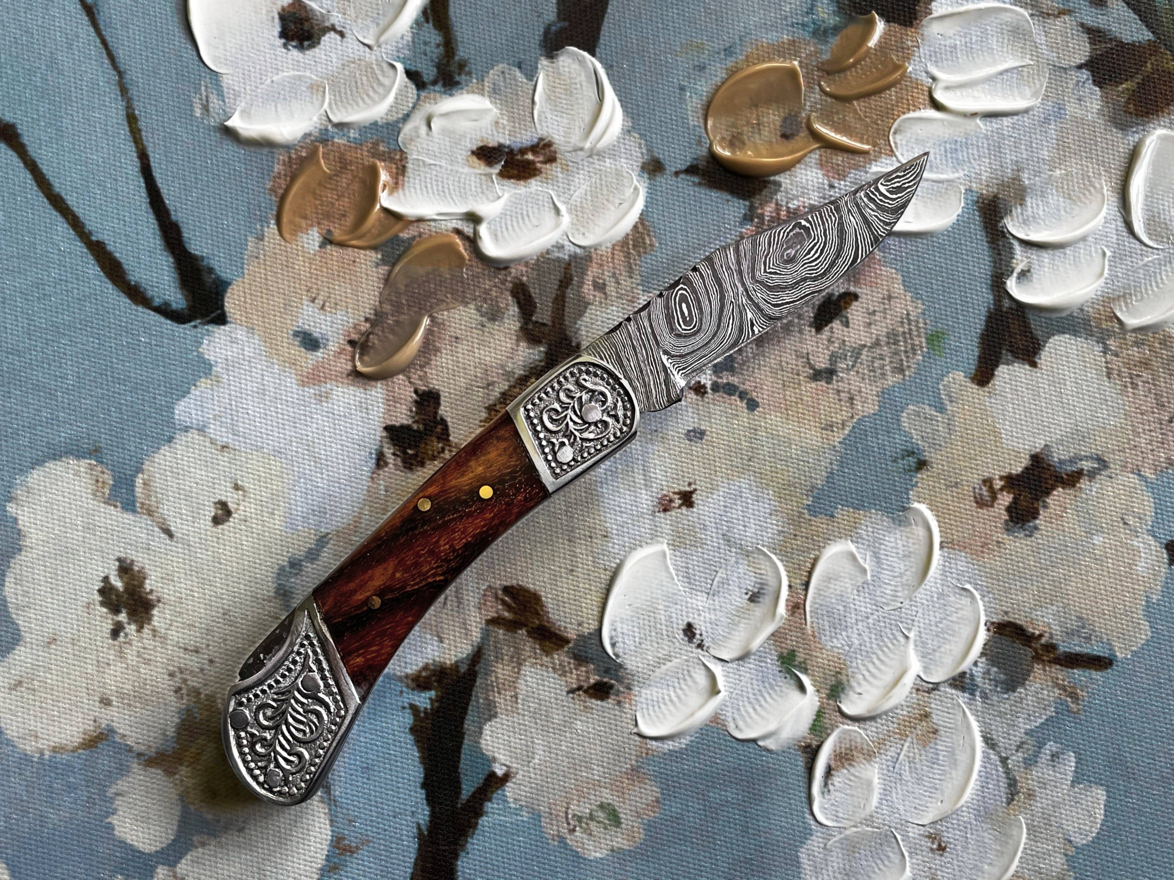 The Knife: Pocket Knife Walnut Wood 6.5'' Damascus Steel, featuring a patterned blade and decorative walnut handle, rests on a textured floral painting.