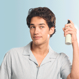 A young man with wavy dark hair, dressed in a light blue shirt, confidently holds a bottle of KITSCH Spray: Texturizing Sea Salt Hair Spray with Rosemary & Biotin (6 fl oz) near his head, gazing to the side against a soft blue background.