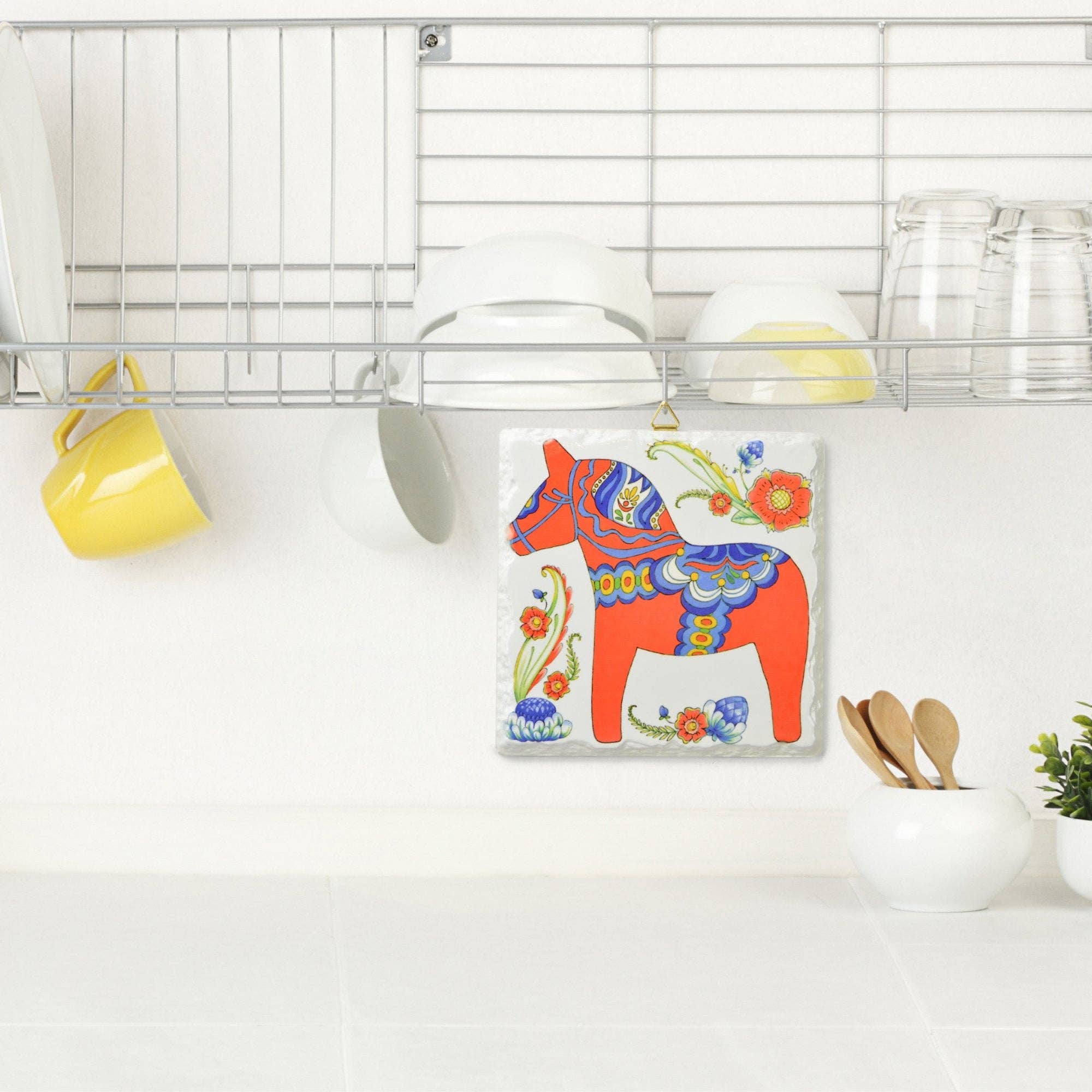 The Red Swedish Dala Horse Ceramic Deluxe Plaque, a 6x6 beveled tile with hanger and cork backing, decorates a white kitchen wall beneath a metal dish rack filled with cups, bowls, and glasses.