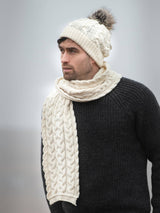 A man in a black sweater, cream cable knit scarf, and the Chunky Cable Hat - Natural (One Size), a merino wool hat with pom-pom made in Ireland, looks to the side against a neutral background.