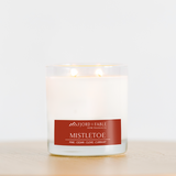 A white Candle: Mistletoe Premium Soy Candle by Fjord & Fable, 4oz jar with two wicks and 20+ hour burn, sits on wood. Notes of pine, cedar, clove, and currant evoke the festive spirit this season in every glowing moment.