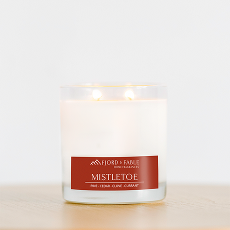 A white Candle: Mistletoe Premium Soy Candle by Fjord & Fable, 4oz jar with two wicks and 20+ hour burn, sits on wood. Notes of pine, cedar, clove, and currant evoke the festive spirit this season in every glowing moment.