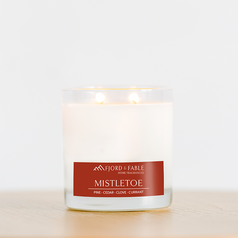 A 4oz Mistletoe Premium Soy Candle from Fjord & Fable, crafted with vegan soy wax and infused with the fragrances of pine, cedar, clove, and currant, is lit and placed on a wooden surface.