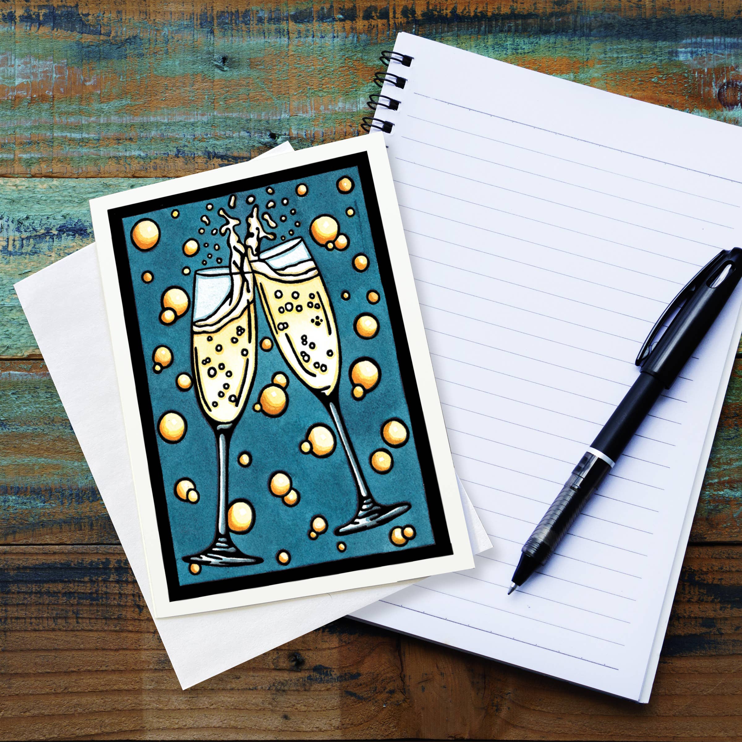 A "Celebration Champagne" greeting card, showcasing an illustration of two clinking champagne glasses with bubbles in the style of Sarah Angst Art's linocut prints, is placed on an envelope next to a lined notebook and black pen on a wooden surface.